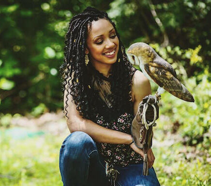 Corina Newsome kneels is a grassy field holding a barn owl on her arm